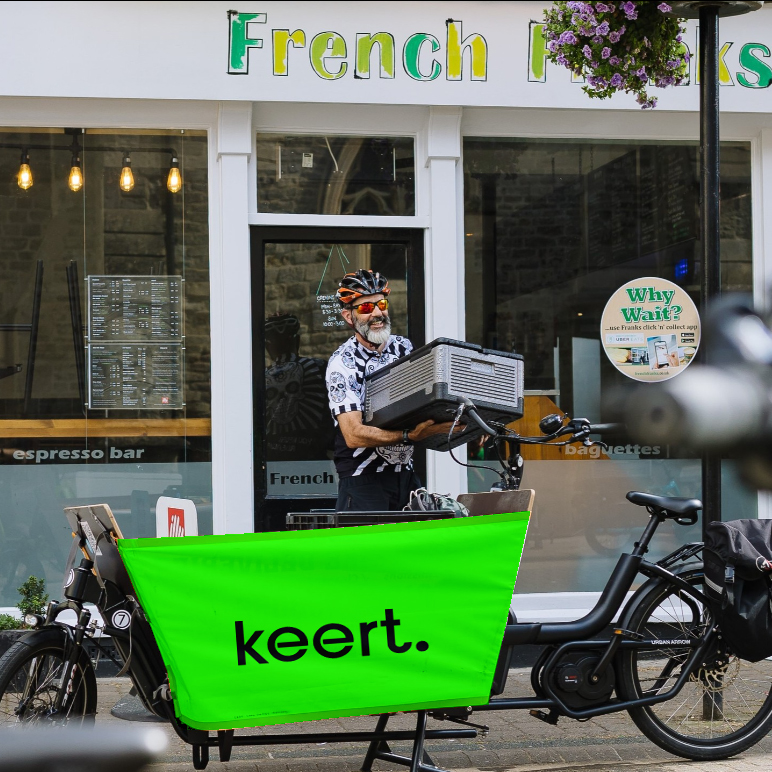 A photo of a cargo bike in the foreground and an white male cyclist wearing sunglasses and a helmet is just leaving a cafe holding a grey plastic basket of baked goods in the background. The cargo bike is branded in neon green with black letting spelling out Keert.