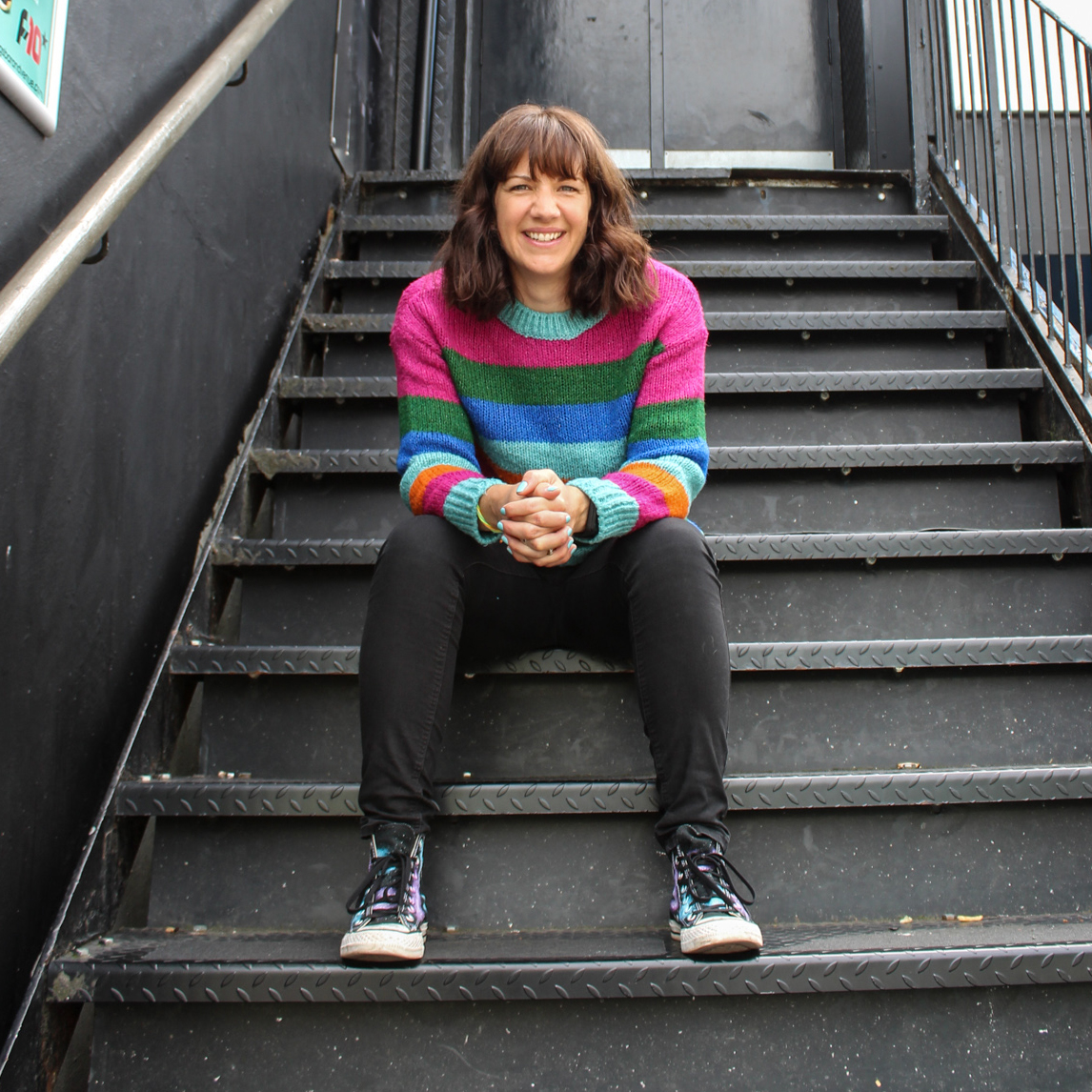 Sara wearing a colourful stripey jumper sat on black metal stairs